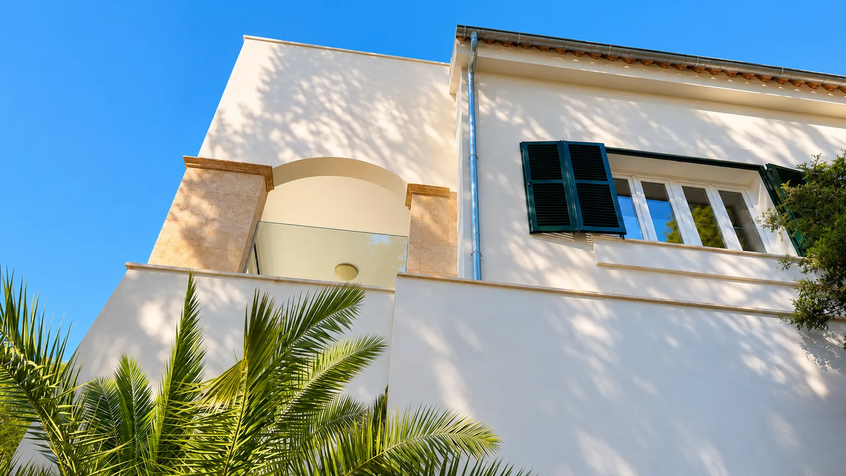Sunlit Mediterranean house façade with green shutters, stone arch, palm leaves and tree shadows.